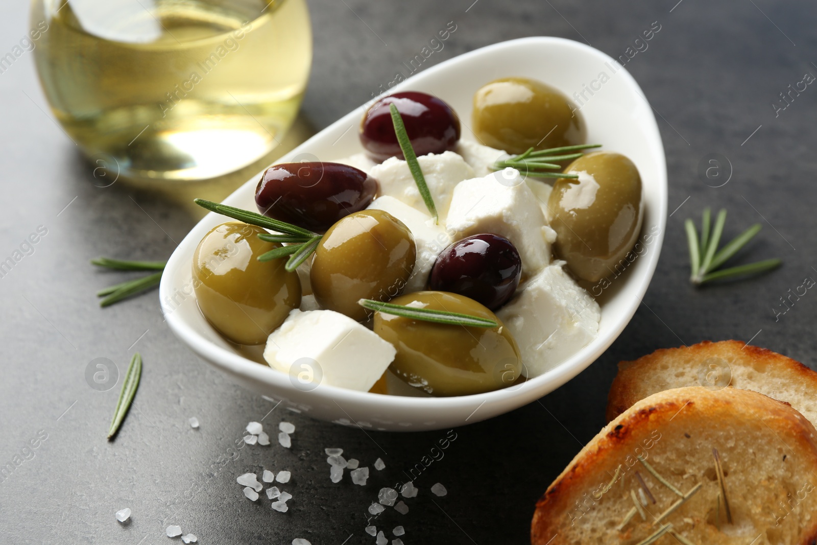 Photo of Marinated olives with feta cheese, bread pieces, oil, salt and rosemary on grey table, closeup