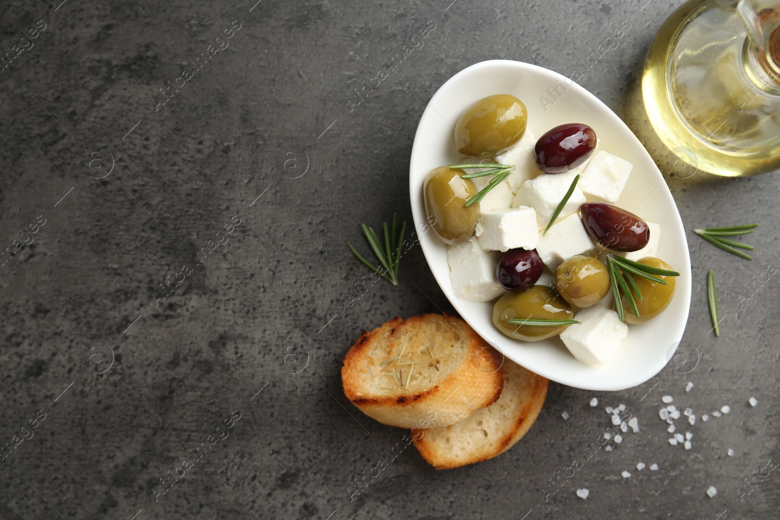 Photo of Marinated olives with feta cheese, bread pieces, oil, salt and rosemary on grey table, flat lay. Space for text