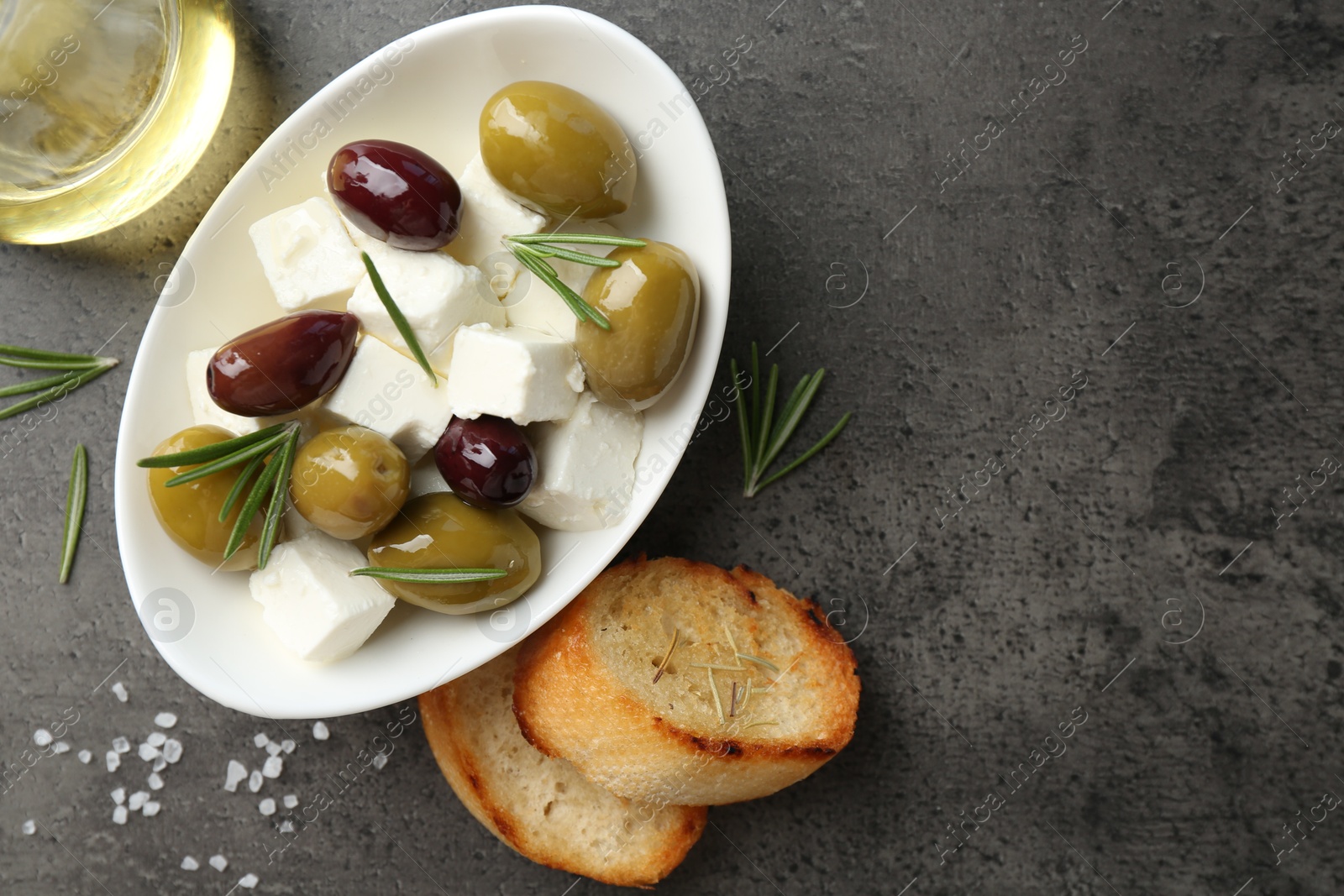 Photo of Marinated olives with feta cheese, bread pieces, oil, salt and rosemary on grey table, flat lay. Space for text