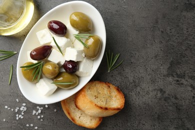 Photo of Marinated olives with feta cheese, bread pieces, oil, salt and rosemary on grey table, flat lay. Space for text