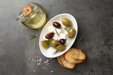 Photo of Marinated olives with feta cheese, bread pieces, oil, salt and rosemary on grey table, flat lay