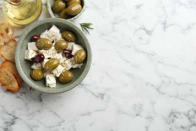 Photo of Marinated olives with feta cheese, bread pieces, oil and rosemary on white marble table, flat lay. Space for text