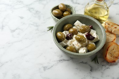 Photo of Marinated olives with feta cheese, bread pieces, oil and rosemary on white marble table. Space for text