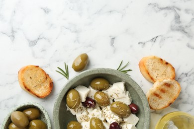 Photo of Marinated olives with feta cheese, bread pieces, oil and rosemary on white marble table, flat lay. Space for text