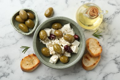 Photo of Marinated olives with feta cheese, bread pieces, oil and rosemary on white marble table, flat lay