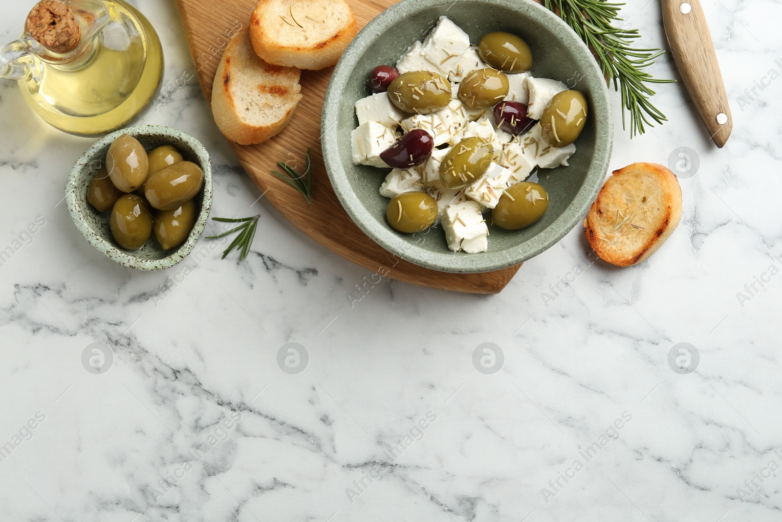 Photo of Marinated olives with feta cheese, bread pieces, oil and rosemary on white marble table, flat lay. Space for text