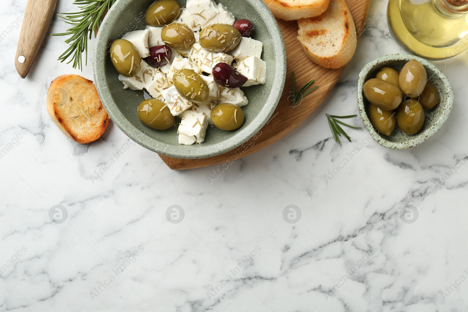 Photo of Marinated olives with feta cheese, bread pieces, oil and rosemary on white marble table, flat lay. Space for text