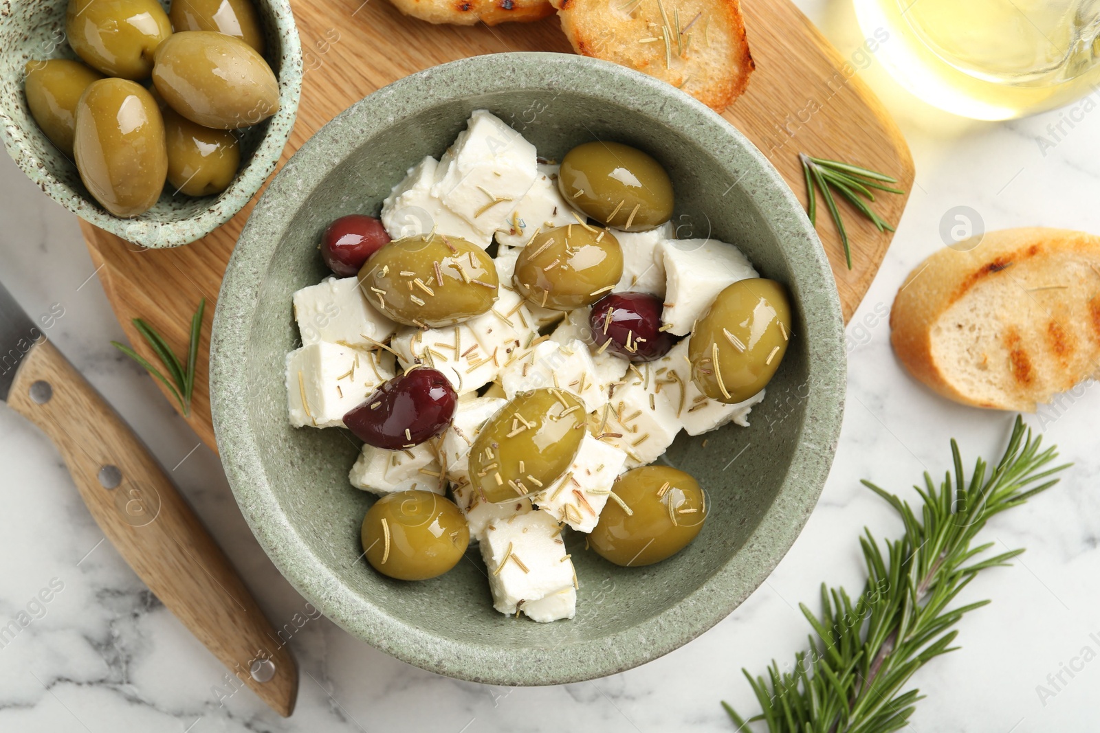 Photo of Marinated olives with feta cheese, bread pieces, oil and rosemary on white marble table, flat lay