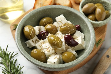 Photo of Marinated olives with feta cheese, bread pieces, oil and rosemary on white marble table, closeup