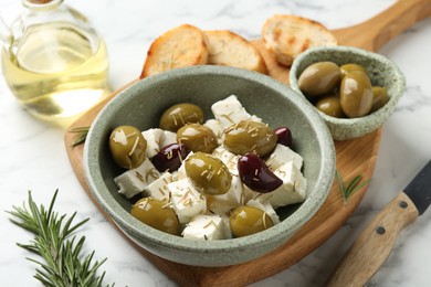 Photo of Marinated olives with feta cheese, bread pieces, oil and rosemary on white marble table, closeup