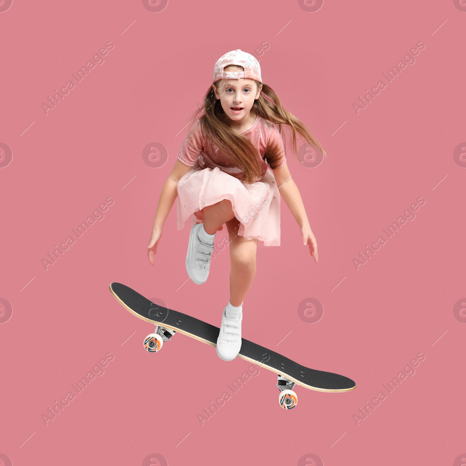 Image of Cute little girl with skateboard in air on pink background. Performing trick