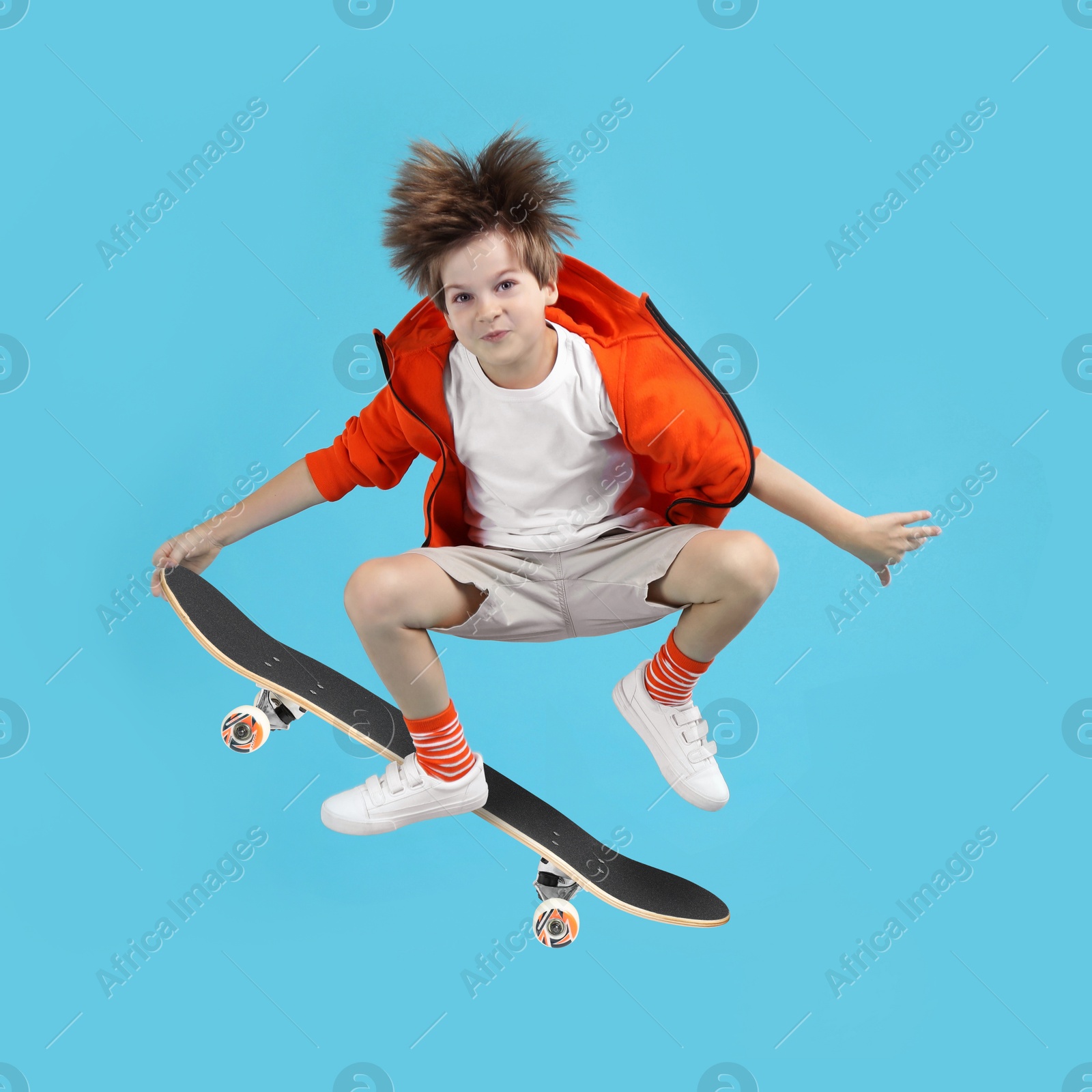 Image of Cute little boy with skateboard in air on light blue background. Performing trick