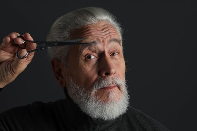 Photo of Bearded senior man trimming eyebrow with scissors on dark grey background