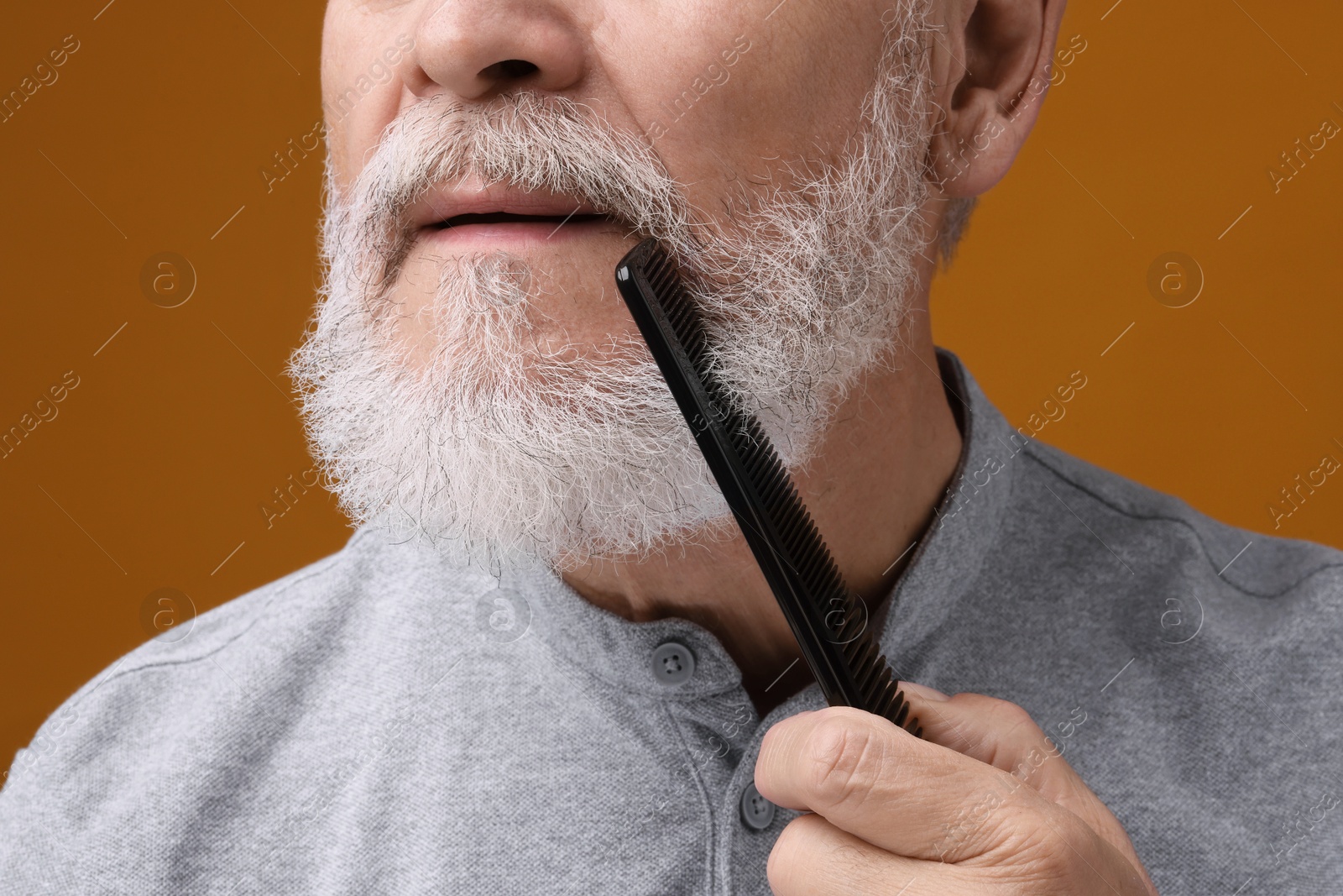 Photo of Senior man combing beard on orange background, closeup