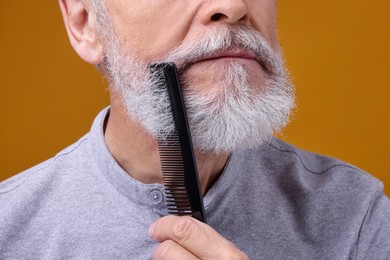 Photo of Senior man combing beard on orange background, closeup