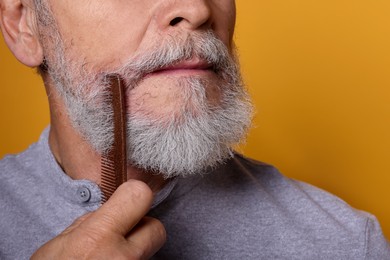 Photo of Senior man combing beard on orange background, closeup