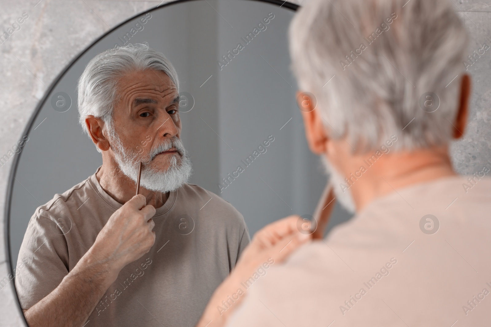 Photo of Senior man combing beard near mirror in bathroom