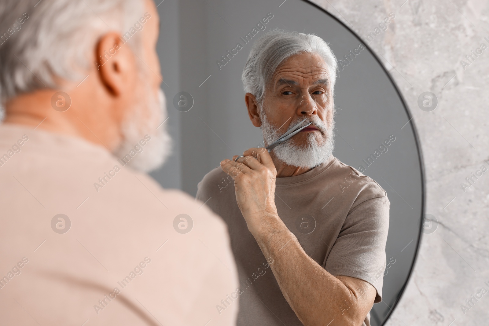 Photo of Senior man trimming beard with scissors near mirror in bathroom