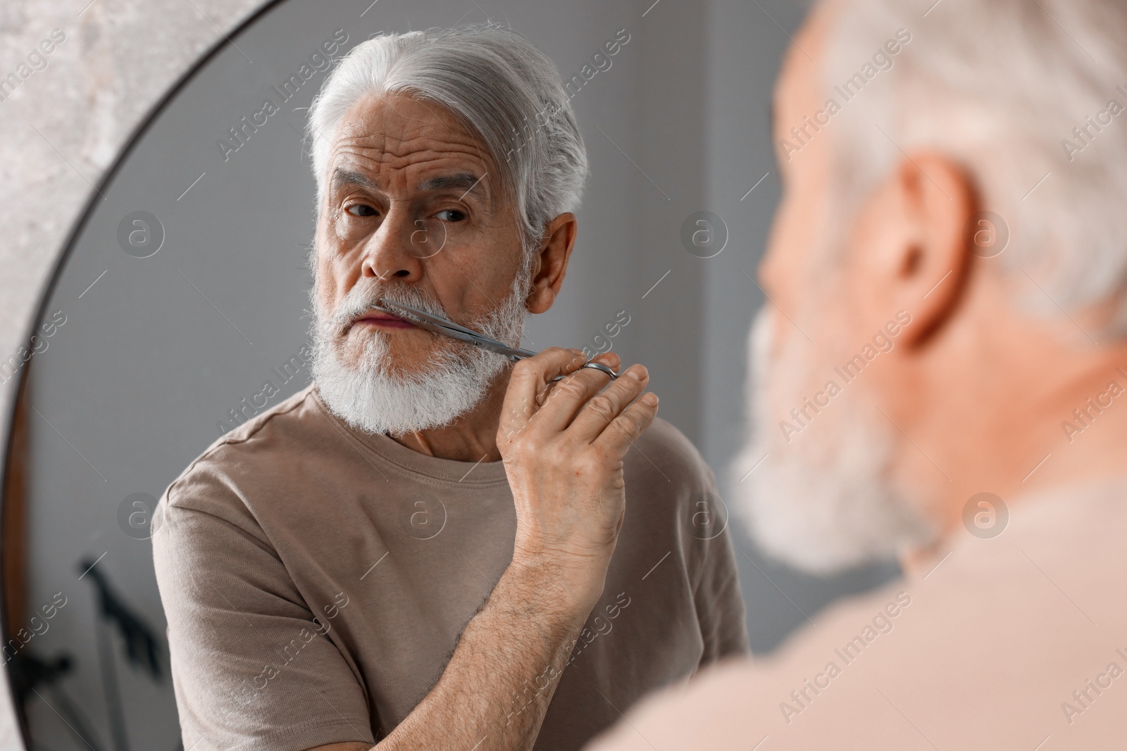 Photo of Senior man trimming beard with scissors near mirror in bathroom