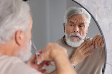 Photo of Senior man trimming beard with scissors near mirror in bathroom