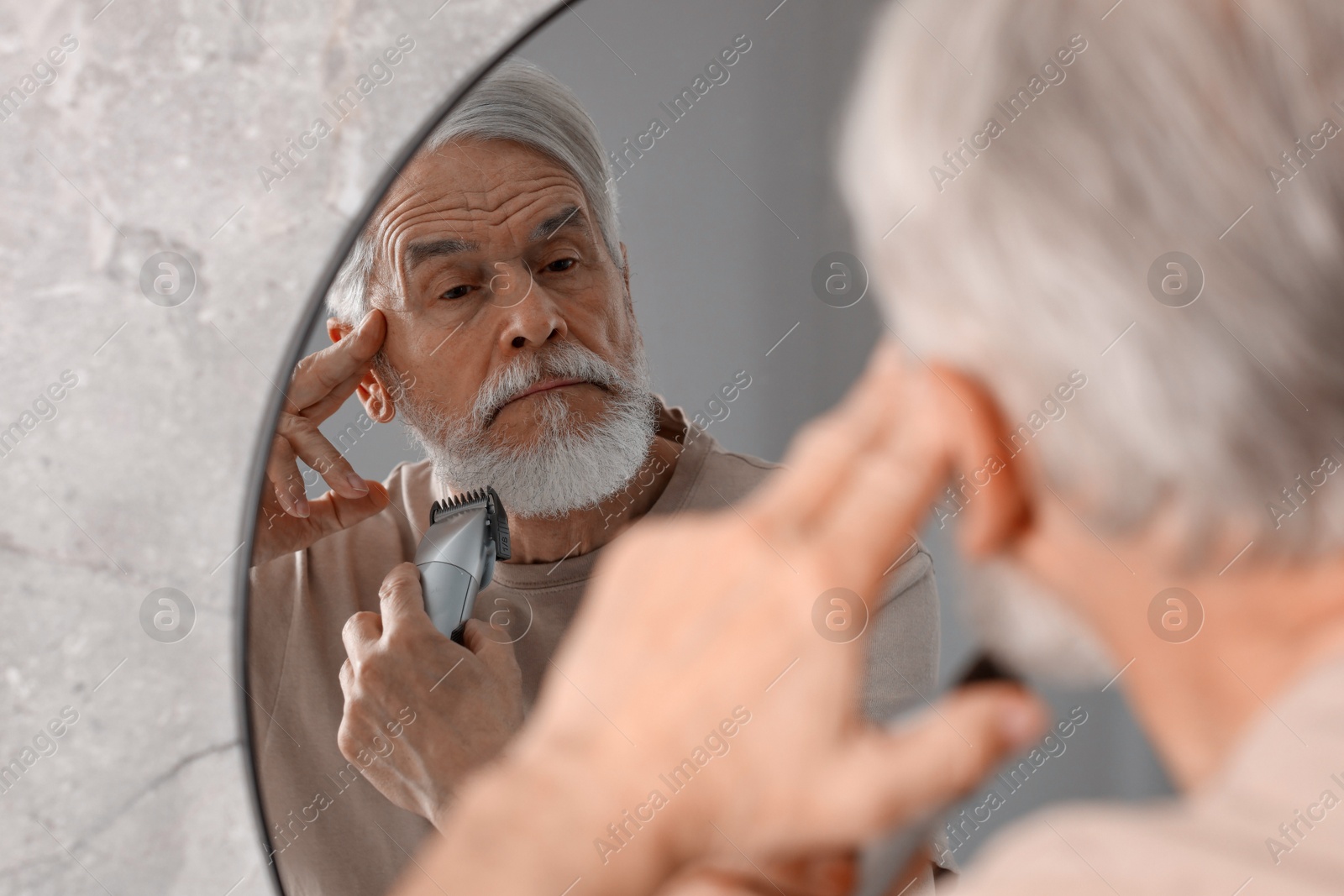 Photo of Senior man trimming beard near mirror in bathroom