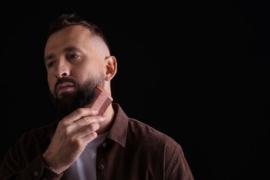 Photo of Handsome man combing beard on black background, space for text