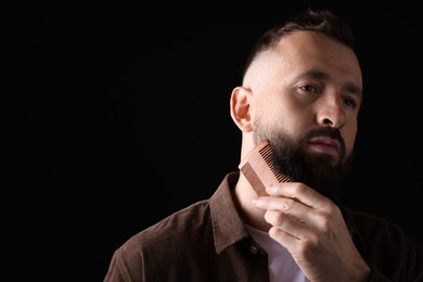 Photo of Handsome man combing beard on black background, space for text