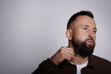 Handsome man combing beard on grey background, space for text