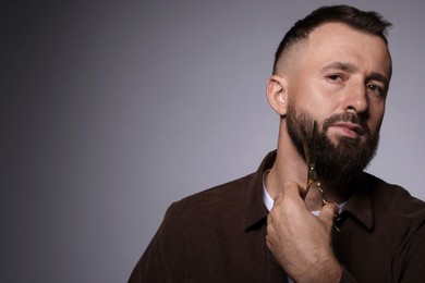 Man trimming beard with scissors on grey background, space for text