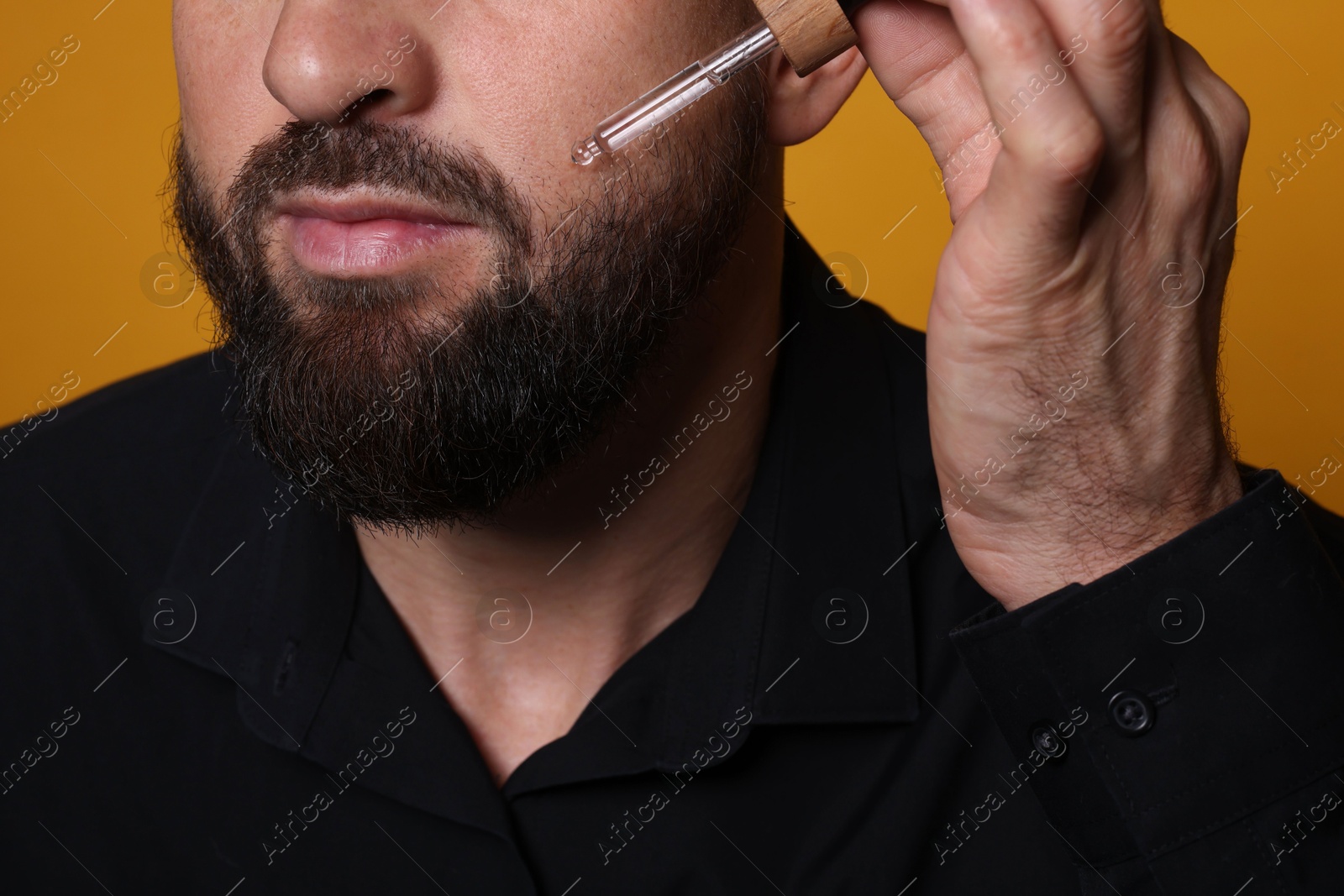 Photo of Man applying serum onto his beard on orange background, closeup