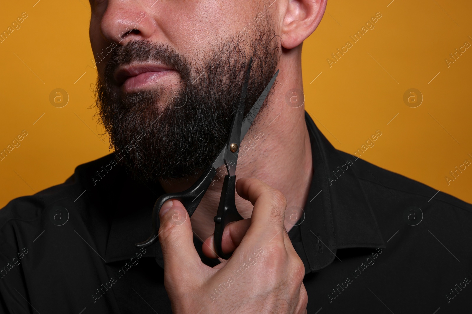 Photo of Man trimming beard with scissors on orange background, closeup