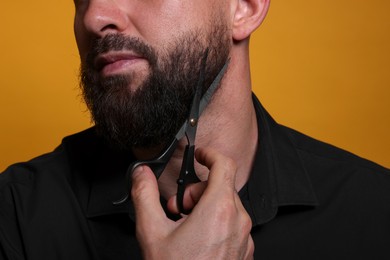 Photo of Man trimming beard with scissors on orange background, closeup