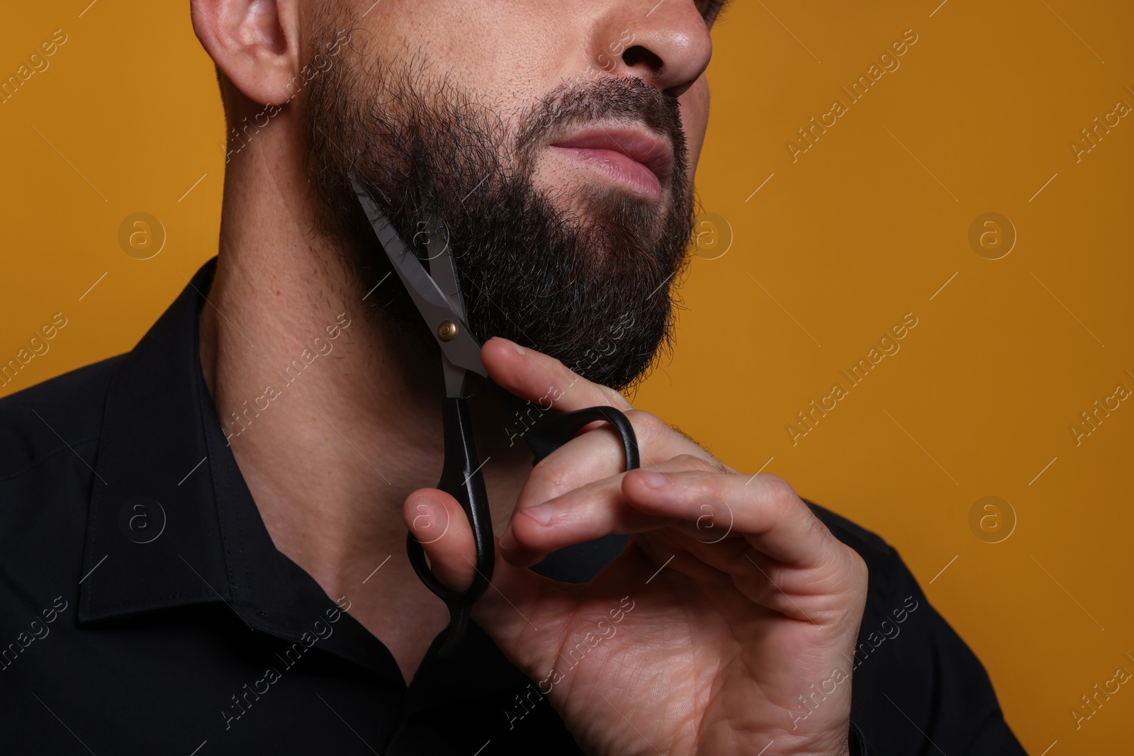 Photo of Man trimming beard with scissors on orange background, closeup