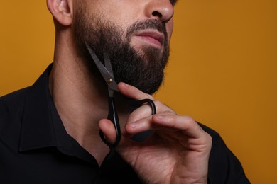 Photo of Man trimming beard with scissors on orange background, closeup