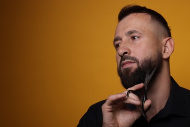 Man trimming beard with scissors on orange background, space for text