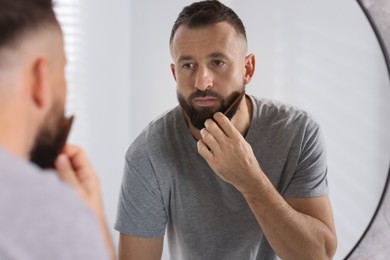 Photo of Man combing beard near mirror in bathroom