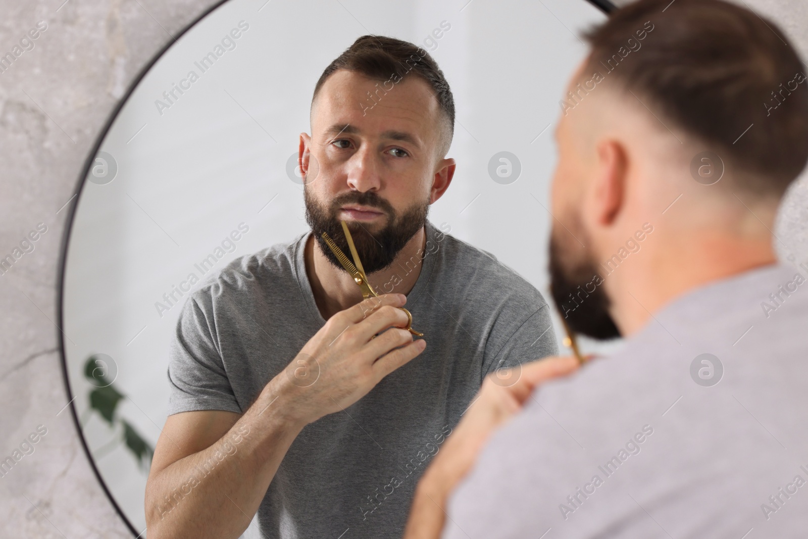 Photo of Man trimming beard with scissors near mirror in bathroom