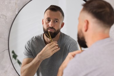 Photo of Man trimming beard with scissors near mirror in bathroom