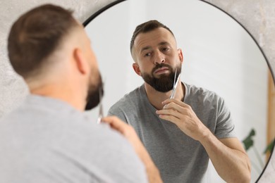 Photo of Man trimming beard with scissors near mirror in bathroom