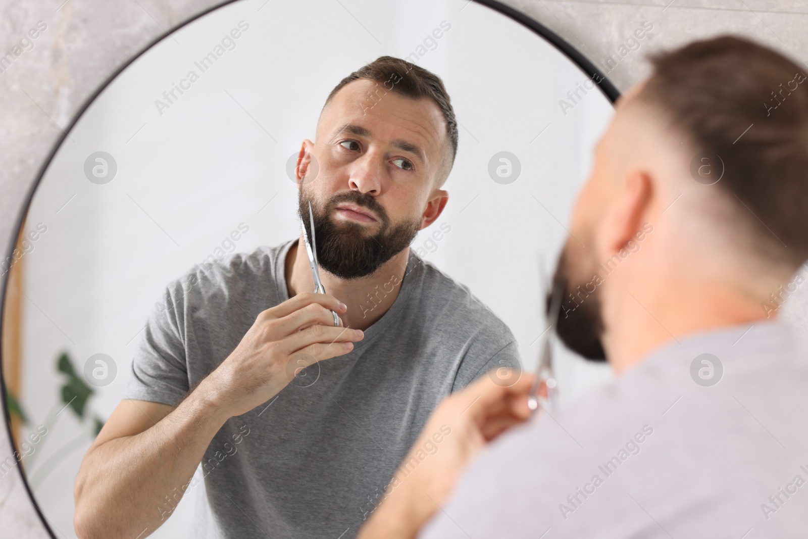 Photo of Man trimming beard with scissors near mirror in bathroom