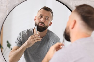 Photo of Man trimming beard with scissors near mirror in bathroom