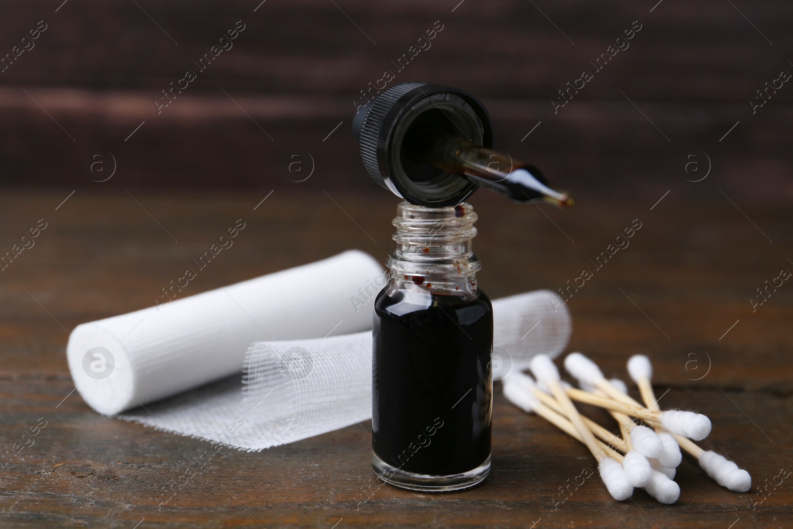 Photo of Bottle of iodine with dropper, cotton swabs and bandage on wooden table, closeup