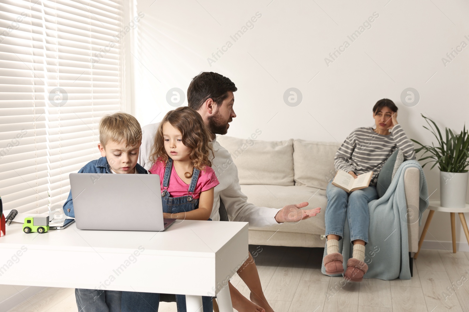 Photo of Father and children at desk with laptop while their overwhelmed mother on sofa indoors