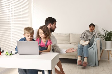 Photo of Father and children at desk with laptop while their overwhelmed mother on sofa indoors