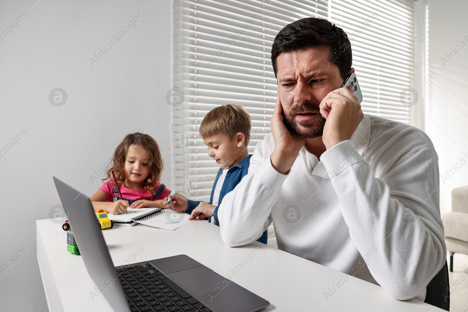 Photo of Naughty children disturbing overwhelmed father while he talking on smartphone at table indoors