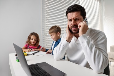 Photo of Naughty children disturbing overwhelmed father while he talking on smartphone at table indoors