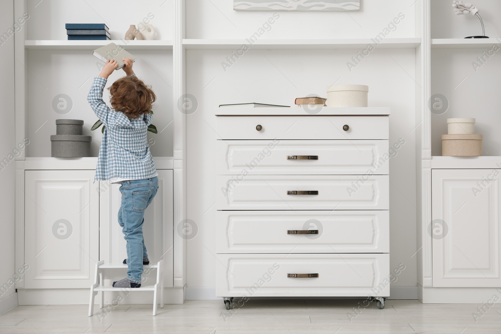 Photo of Little boy with book standing on step stool near shelving unit at home, back view