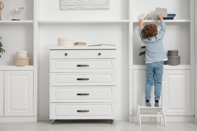 Photo of Little boy standing on step stool and reaching for book at home, back view