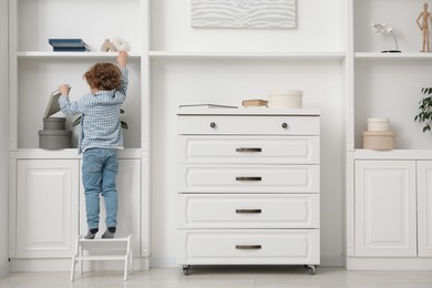 Photo of Little boy with book standing on step stool near shelving unit at home, back view