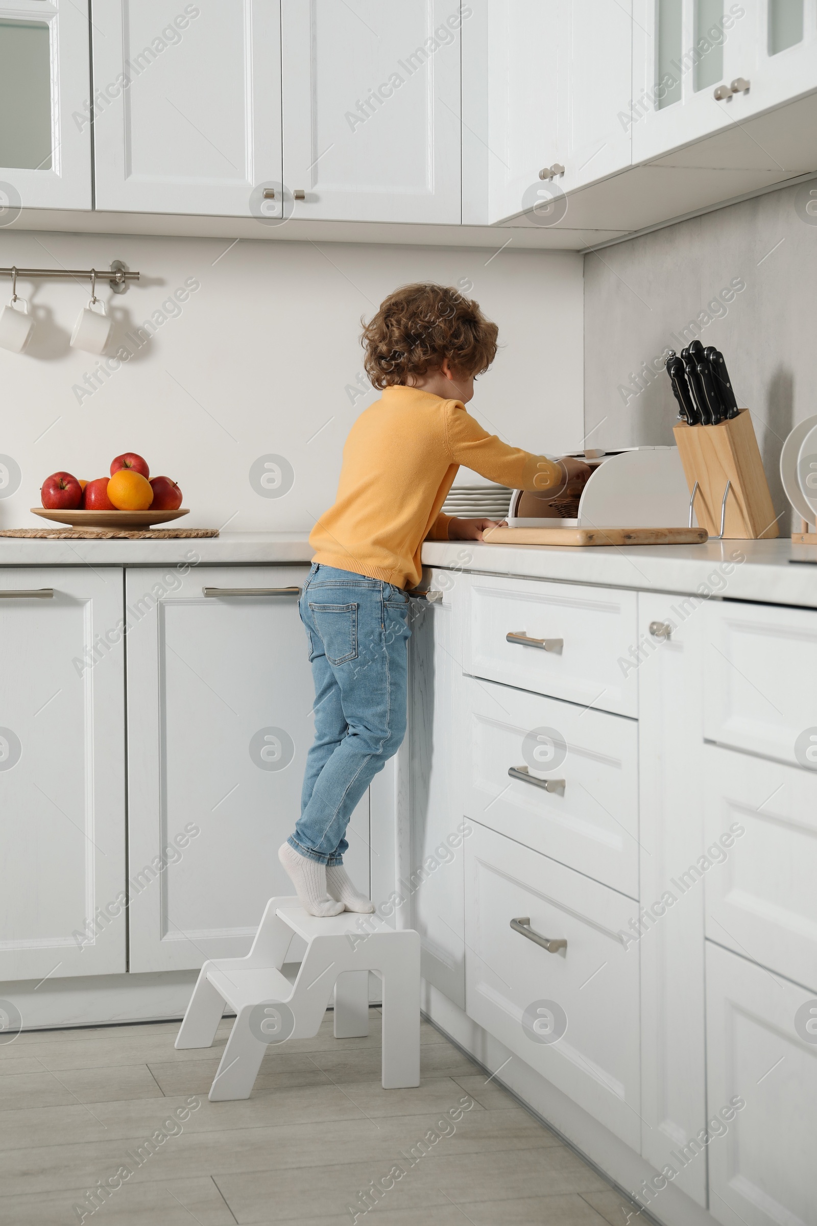 Photo of Little boy standing on step stool and reaching towards bread box in kitchen